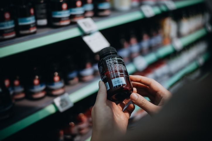 Over the shoulder view of woman browsing through medical products and reading the label on a bottle of medicine in front of the shelves in a pharmacy. Healthcare, medication and people concept