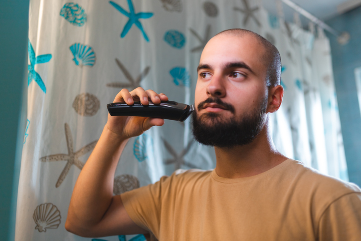 Close-up of a young bald man trimming his beard in front of the mirror