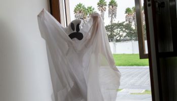 A funny little cute girl wearing a ghost costume having fun and dancing in Halloween's season. She's standing in front of the main door asking for candies in a Halloween afternoon day.