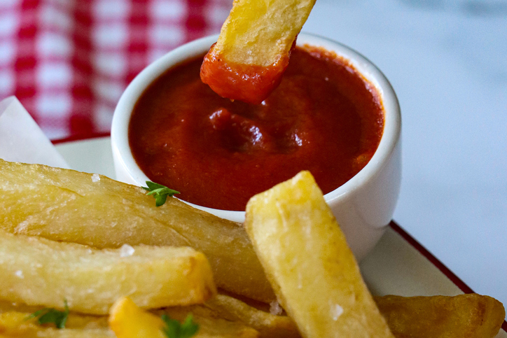 Full frame image of greaseproof parchment paper lined enamel bowl of freshly cooked crispy homemade French fries garnished with chopped parsley and salt, chip dipped in ramekin of tomato ketchup, red and white gingham checked cloth background