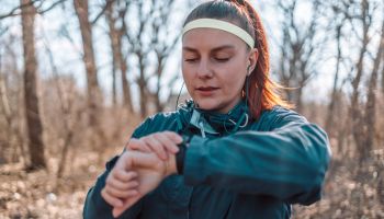 Sport watch run woman checking smartwatch tracker. Trail running runner girl looking at heart rate monitor smart watch in forest wearing jacket sportswear. Female athlete jogger training in woods.