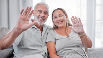 Elderly couple, relax and smile in hello for introduction, waving hands or video call on living room sofa at home. Portrait of happy senior man and woman smiling in happiness for communication