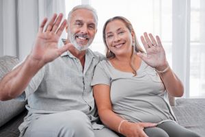 Elderly couple, relax and smile in hello for introduction, waving hands or video call on living room sofa at home. Portrait of happy senior man and woman smiling in happiness for communication