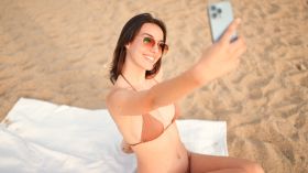 Clouse-up, slim beautiful brown-haired woman with long hair uses mobile phone. Smiling girl wearing swimsuit makes selfie sits on the sand on the beach.