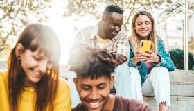 Multicultural millennial people using smart mobile phone device sitting outdoors - Happy teenagers having fun watching reel video on social media platform - Trendy technology and youth culture concept