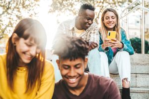 Multicultural millennial people using smart mobile phone device sitting outdoors - Happy teenagers having fun watching reel video on social media platform - Trendy technology and youth culture concept