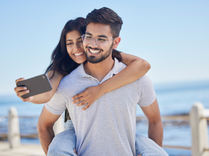 Phone selfie, ocean and couple hug, bond and enjoy time together for peace, freedom or romantic date. Sea beach, memory photo and man piggyback woman on fun travel holiday in Rio de Janeiro Brazil