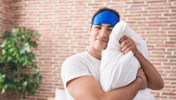 Young hispanic man wearing sleep mask hugging pillow at bedroom