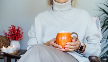 Close up pumpkin shaped cup of hot coffee drink with marshmallow in female hands with fall mood decor for hygge home on background. Cozy autumn beverage for Thanksgiving or Halloween holidays