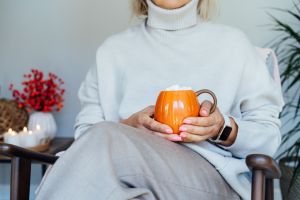Close up pumpkin shaped cup of hot coffee drink with marshmallow in female hands with fall mood decor for hygge home on background. Cozy autumn beverage for Thanksgiving or Halloween holidays