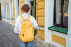 A teenage girl with a yellow school backpack walks along the street along the walls of the school.