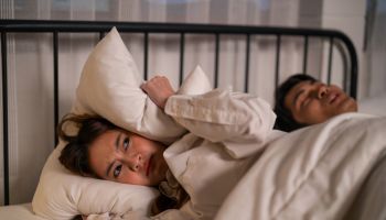 Asian woman feel frustrated from husband snoring while sleep at night. Attractive insomnia angry wife lying down on bed, cannot sleep due to noise and put cozy blanket on her ears in bedroom at home.