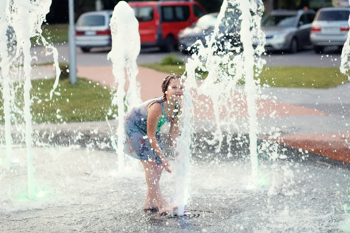 Little cute funny girl child bathes on a hot summer day in a public city fountain. The child runs, plays among the jets and splashes of water
